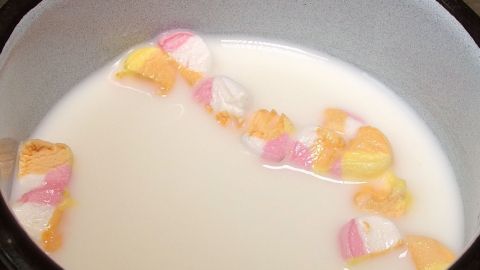 Eine Tasse heiße Schokolade mit bunten Marshmallows schwimmt in einer weißen, cremigen Flüssigkeit.