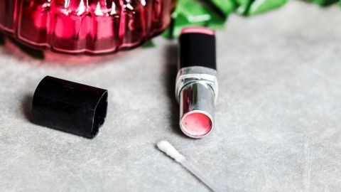 Ein Lippenstift liegt offen neben einem eleganten Parfümflakon, eingerahmt von grünen Blättern auf einer neutralen Oberfläche.