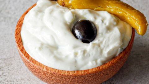 Eine Holschüssel enthält cremiges Zaziki, garniert mit einer schwarzen Olive und einer grünen Chili.