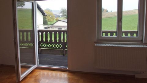 Ein offenes Fenster ermöglicht frische Luft in einem hellen Raum mit Holzfußboden und Blick auf eine grüne Landschaft.