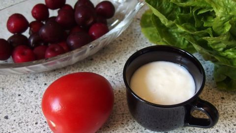 Eine rote Tomate liegt neben einer schwarzen Tasse mit einer Creme, umgeben von frischem Salat und Trauben auf einer Arbeitsfläche.