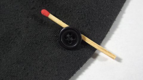 Während des Annähens unterlegt man den Knopf mit einem Streichholz, dadurch bekommt der Knopf einen "Hals" und lässt sich nun mühelos durch das Knopfloch schieben. Ein schwarzer Stoff mit einem Knopf und einem roten Streichholz, das quer über den Stoff gelegt ist.
