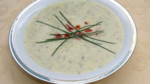 Eine cremige Zucchini-Cremesuppe in einer weißen Schüssel, verziert mit Kräutern und roten Punkten, auf einem eleganten Tisch.