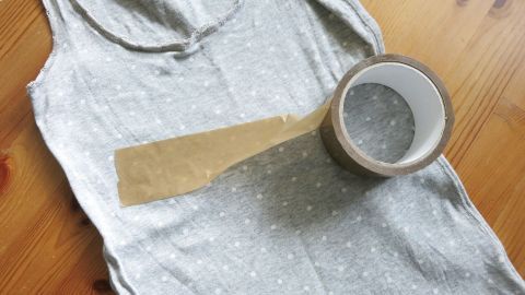 Schwarze Fussel auf Kleidung entfernt man leicht und erfolgreich mit Paketband. Probier's aus. Ein graues Tanktop liegt auf einem Holztisch, neben einer Rolle Klebeband, um schwarze Fusseln zu entfernen.