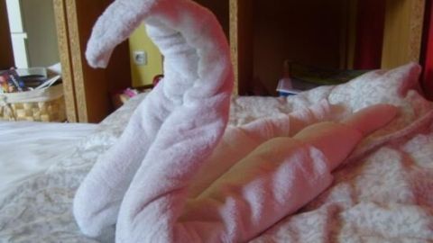 Ein Handtuchtier in Schwanform liegt dekorativ auf einem Bett vor einem Regal mit Körben und Büchern.
