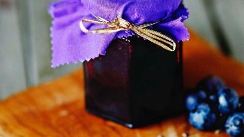 Ein Glas Blaubeer-Vanillemarmelade mit Lavendel steht auf einem Holzbrett, umgeben von frischen Blaubeeren und getrocknetem Lavendel.