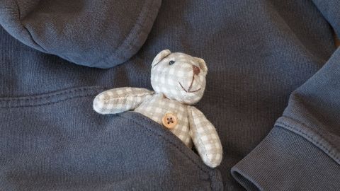 Ein kleiner, gestreifter Teddybär sitzt fröhlich in der Tasche eines alten, grauen Pullis und lächelt charmant.