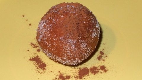 Ein Quarkbällchen liegt auf einem gelben Teller, umgeben von Kakaopulver, das einen süßen Snack andeutet.