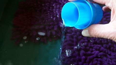 Eine Hand schüttet Hygienespüler aus einer blauen Flasche in ein Wasserbad mit einer lila Textiloberfläche.