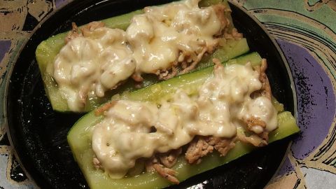 Zucchinihälften sind gefüllt mit Hack und überbacken, serviert auf einem dunklen Teller mit buntem Hintergrund.