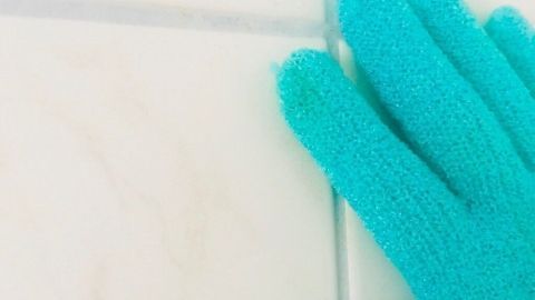 Ein grüner Peeling-Handschuh reinigt Fliesenfugen und entfernt Schmutz in einem hellen Badezimmer.