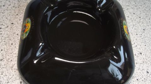 Ein schwarzer Aschenbecher liegt auf einer hellen, gemusterten Oberfläche und scheint unbenutzt zu sein.