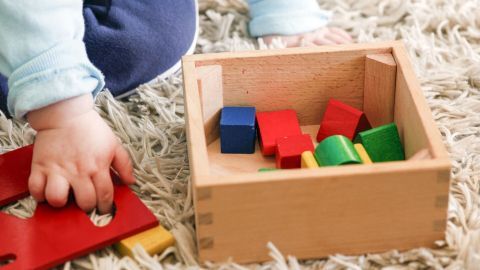 Ein Kind greift mit einer Hand nach Bauklötzen, die in einer Holzbox auf einem Teppich verteilt sind.