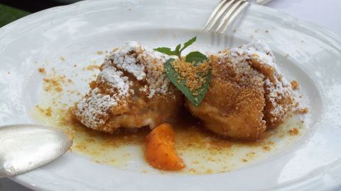 Der Teig für die Marillenknödel kann bereits am Vortag zubereitet werden, und im Kühlschrank “übernachten“. Zwei Marillenknödel auf einem weißen Teller, bestäubt mit Puderzucker, liegen in einer süßen Soße und frischen Kräutern.
