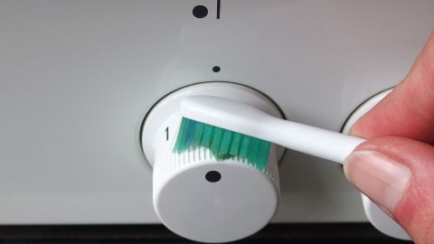 Die Schalter eines Elektroherds lassen sich mithilfe von Glasreiniger und einer Zahnbürste reinigen. Ein Handschuh hält eine Zahnbürste, die gerade einen Elektroherd-Schalter reinigt, um Schmutz zu entfernen.