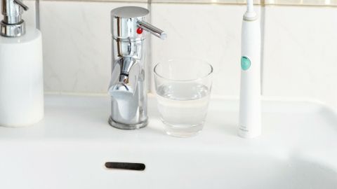Blitzblankes Zahnputzglas mithilfe von Salz. Jetzt den Tipp lesen und ausprobieren. Ein elektrischer Zahnbürste steht neben einem Glas Wasser auf einem Waschbecken, umgeben von Pflanzen in goldenen Töpfen.