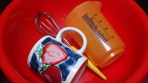 Eine rote Spülschüssel enthält einen Schneebesen, einen Messbecher und eine Tasse mit Erdbeeren, alle im Wasser.