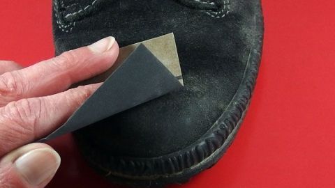 Eine Hand reinigt einen schwarzen Wildlederschuh mit einem speziellen Werkzeug auf einer roten Oberfläche.