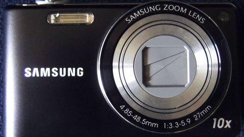 Eine schwarze Digitalkamera von Samsung liegt mit dem Objektiv nach oben auf einem blauen Untergrund. Die Kamera ist bereit für Aufnahmen.