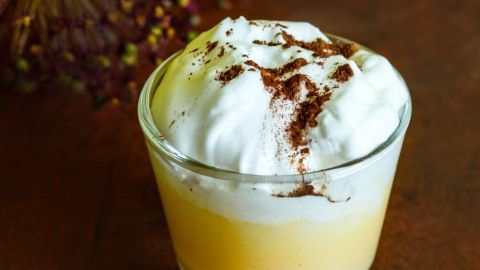 Ein Glas Eierlikör ohne kochen, garniert mit Sahne und Kakaopulver, steht vor einer dekorativen, unscharfen Pflanze.