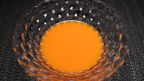 Ein tiefes Schüsselchen mit leuchtend orangefarbigem Saft steht auf einer dunklen Oberfläche und reflektiert sanft das Licht.