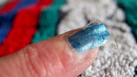 Mithilfe von Tesafilm ausprobieren und sicher gehen, welche Nagellackfarbe einem am besten gefällt. Ein mit bläulichem Nagellack lackierter Fingernagel zeigt einen glänzenden Effekt und hebt sich vor einem bunten Hintergrund ab.