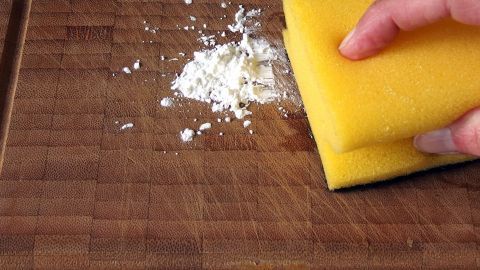 Ein gelber Schwamm reinigt ein Holzbrett, während sich ein wenig Puder darauf verteilt. Es ist eine saubere Küchenumgebung.