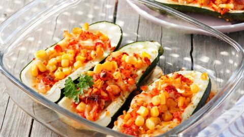 Gefüllte Zucchini liegen in einer Glasform, überbacken mit Gemüse und Käse, auf einem rustikalen Holztisch.