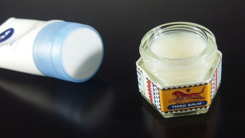 Tigerbalm besser auftragen mit einer Portion Körperlotion. Ein Glas Tigerbalsam steht offen auf einem dunklen Hintergrund, während nebenan eine Tube mit blauer Kappe liegt.
