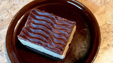 Ein Stück Kuchen mit glatter, dunkler Schokoglasur liegt auf einem runden, braunen Teller. Der Hintergrund ist neutral.