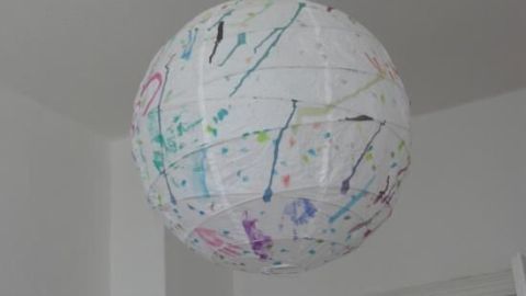 Eine runde Lampe hängt dekorativ von der Decke, verziert mit bunten Klecksen, in einem hellen Raum mit weißer Tür.