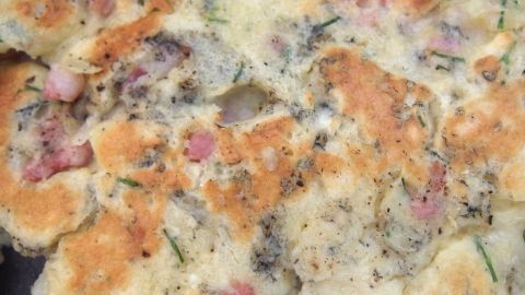 Ein saftiger Eierhannes liegt frisch gebacken auf einem Blech, goldbraun und appetitlich, umgeben von Kräutern und Speckstücken.