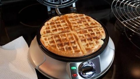 Eine gebackene Waffel liegt im Waffeleisen. Der Metallrost und Papierhandtuch sind daneben; das Gerät zeigt einen grünen Knopf.
