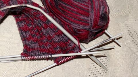 Ein Knäuel rotes Garn aus dem Strümpfestricken liegt neben einem unvollendeten sockenartigen Strickstück auf einer Tischdecke.
