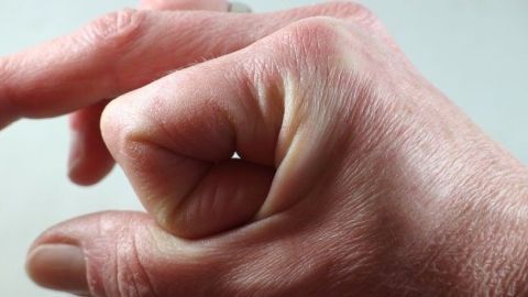 Eine Hand zeigt auf ihren eigenen Handrücken, wobei sie einen Griff simuliert; der Hintergrund ist neutral.