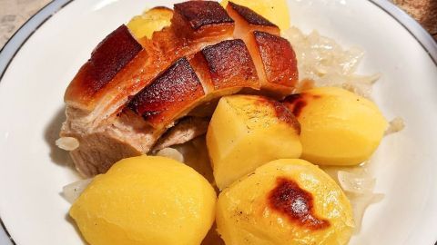 Das Einpinseln des Braten mit Honig kurz vor dem Ende der Garzeit, lässt diesen nochmal knuspriger werden. Goldbraune, knusprige Bratenscheiben liegen auf einem Teller, umgeben von weichgekochten Kartoffeln und Zwiebeln.
