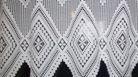 Weißes Gardinenmuster mit aufwendigen Spitzendetails hängt vor einem dunklen Hintergrund und zeigt elegante, transparente Textur.