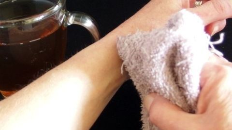 Eine Hand tupft mit einem Tuch auf den Arm, während neben ihr eine Tasse Schwarztee steht, um Juckreiz zu lindern.