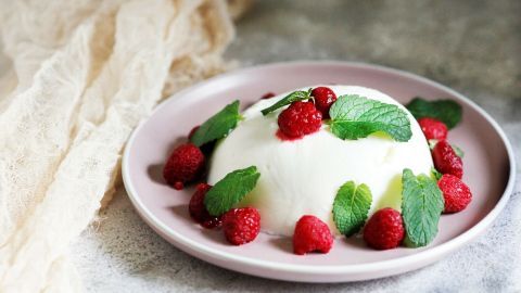 Eine cremige Joghurtbombe mit frischen Himbeeren und Pfefferminzblättern auf einem zarten Teller, umgeben von einem neutralen Hintergrund.