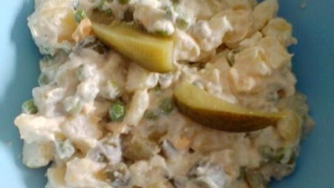 Kartoffelsalat mit Erbsen und Gurkenstücken liegt in einer blauen Schüssel, umgeben von einer cremigen Sauce.