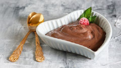 Eine herzförmige Schüssel enthält cremigen Schoko-Quark, garniert mit einer Beere und Minzblatt, auf einer grauen Oberfläche.