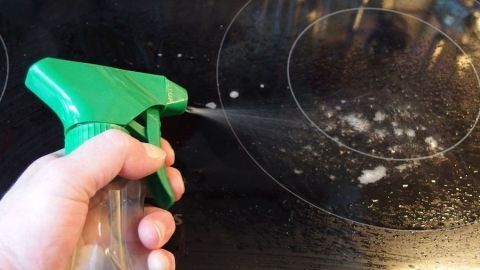 Ein Hand hält einen grünen Sprühkopf, der Fensterreiniger auf ein sauberes Glaskeramikfeld sprüht.