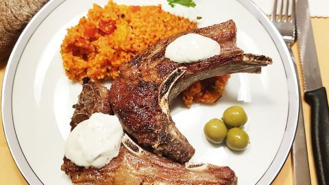 Saftige Lammkeule mit Sauce liegt neben würzigem Couscous und Oliven auf einem Teller, umgeben von Besteck und Brot.