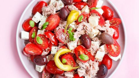 Ein bunter Tomaten-Thunfisch-Salat mit Schafskäse auf einem weißen Teller, umgeben von frischen Kräutern und Oliven.