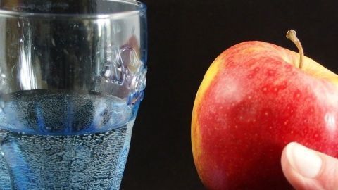 Ein roter Apfel wird von einer Hand gehalten, während ein Glas Wasser daneben steht, auf einem dunklen Hintergrund.