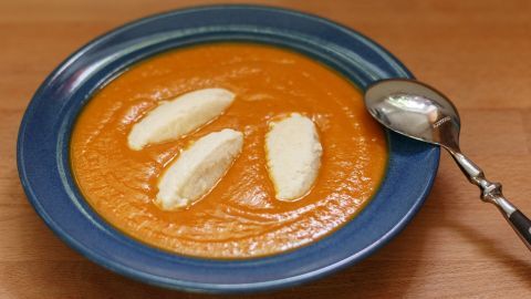 Eine orangefarbene Kürbis-Kokos-Suppe wird in einer blauen Schüssel serviert, garniert mit Sahnehäufchen. Ein Löffel liegt daneben.