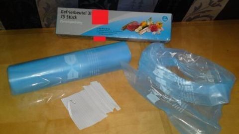 Gefrierbeutel, in Blau aufgerollt, mit Box und Gemüseabbildung. Auf Tisch liegen Streifen. Text: "Gefrierbeutel 3 l, 75 Stück".