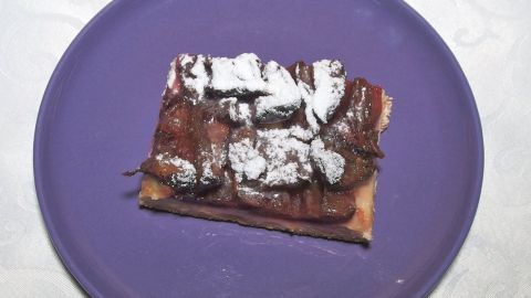 Ein Stück Hefeteiggebäck liegt auf einem lila Teller, bedeckt mit Schokoladensoße und Puderzucker.