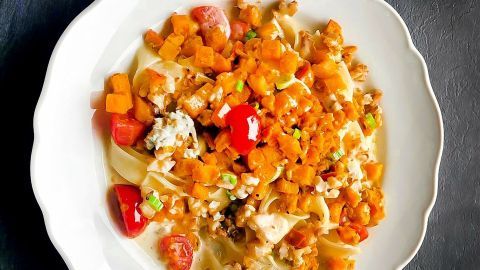 Ein Teller mit überbackenem Kürbis, frischen Tomaten und Nudel-Rahmsoße ruht auf einem dunklen Untergrund.