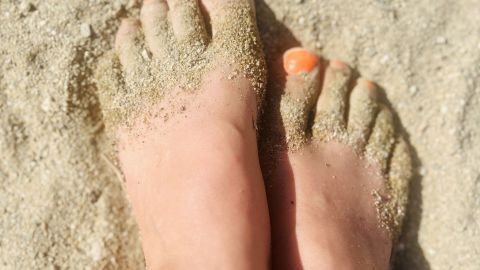 Weiche Füße wie am Strand? Mit diesem Tipp geht das auch zu Hause. Weiche Füße stehen im Sand, während Sonnenlicht darauf scheint und orangefarbener Nagellack leuchtet.